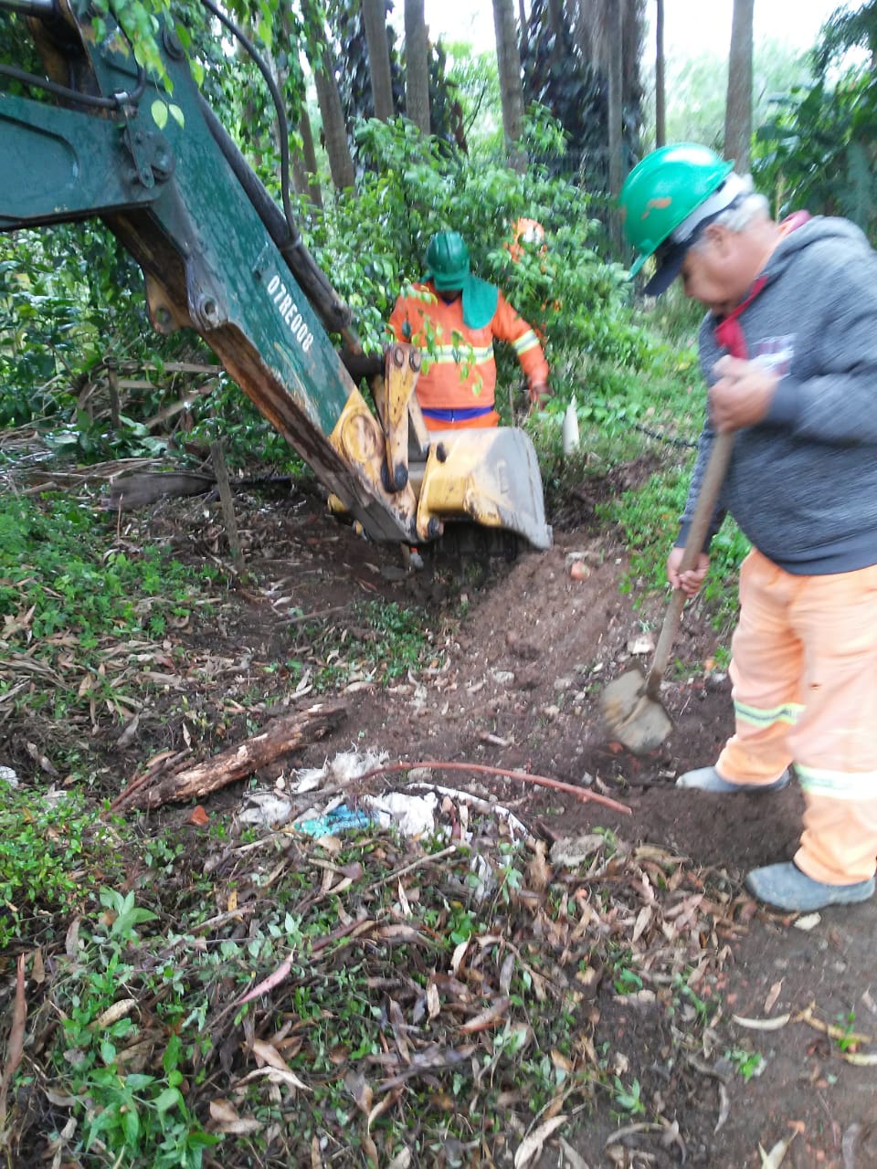 #PraCegoVer - Trabalhadores da Subprefeitura, ajudados por uma escavadeira, limpam o caminho da sangria (escoadouro), em meio a árvores e vegetação.