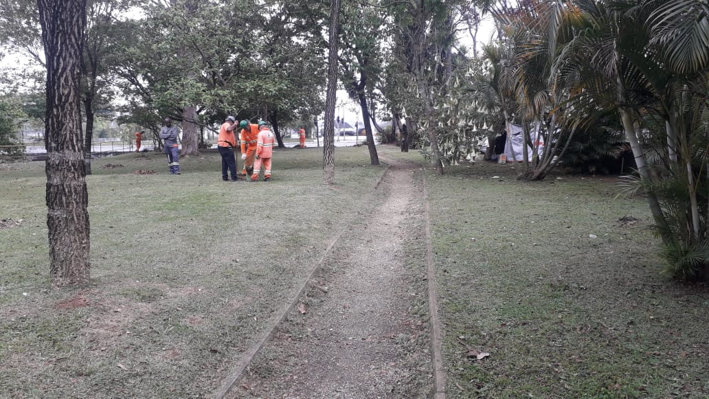 #PraCegoVer - Trabalhadores da Subprefeitura cortam a grama da praça. Há árvores dos dois lados de um caminho de pedestres.