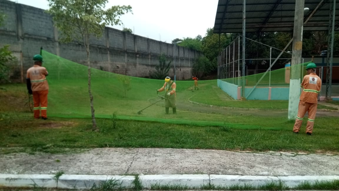 #PraCegoVer - Trabalhadores da Subprefeitura cortam grama perto de uma quadra de esportes. Outros seguram a tela de proteção.