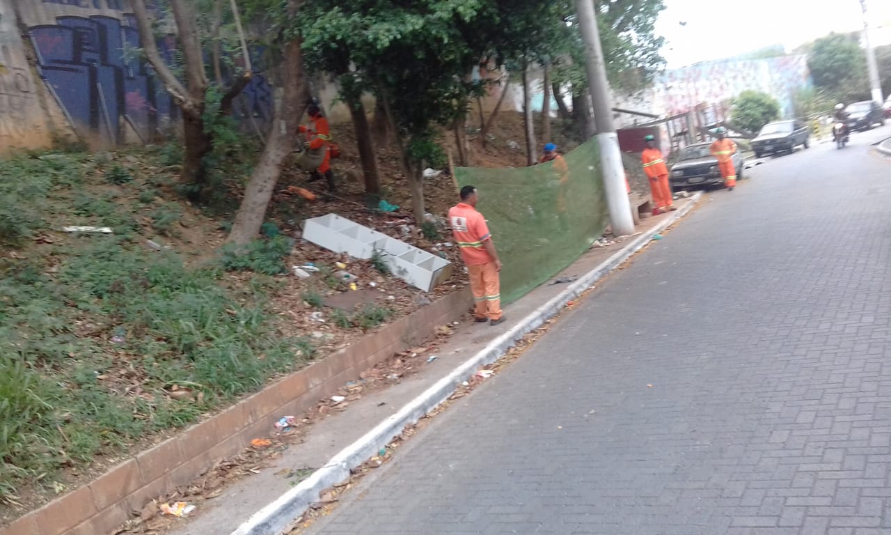 #PraCegoVer - Trabalhadores da Subprefeitura cortam grama em encosta ao lado da via. Outros trabalhadores seguram proteção de tela.