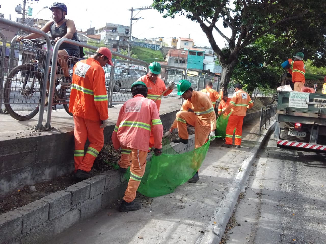 #PraCegoVer - Trabalhadores da Subprefeitura recolhem grama e galhos cortados no canteiro central da via. Há um caminhão, à direita, para onde a grama e os galhos são levados.