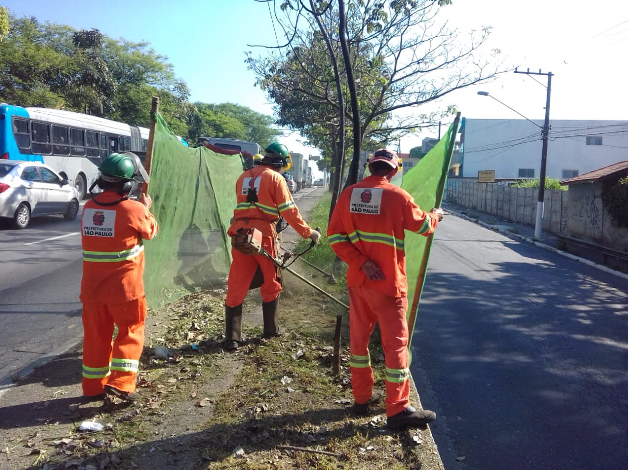 #PraCegoVer - Trabalhadores da Subprefeitura cortam grama no canteiro central. Outros seguram proteções de tela dos dois lados do canteiro.