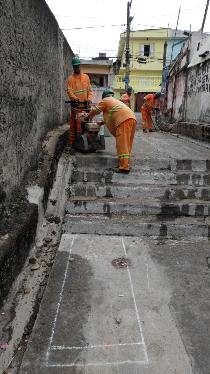 #PraCegoVer - Trabalhadores da Subprefeitura demarcam o local do conserto, outros estão ao fundo, fazendo medições. A viela tem trechos em escadas. Dos dois lados, há muros. No fundo, na saída da viela, um sobrado amarelo limão.