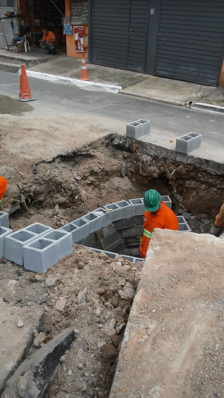 #PraCegoVer - Trabalhador da Subprefeitura está dentro de buraco escavado para alcançar a galeria. Está sendo erguida uma parede interna, com blocos de concreto.