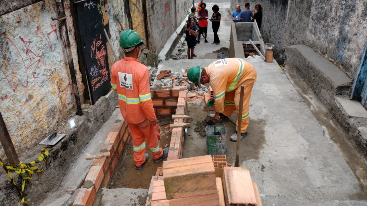 #PraCegoVer - Trabalhadores da Subprefeitura erguem uma caixa com tijolos de cerâmica. Ao fundo, moradores acompanham o trabalho.