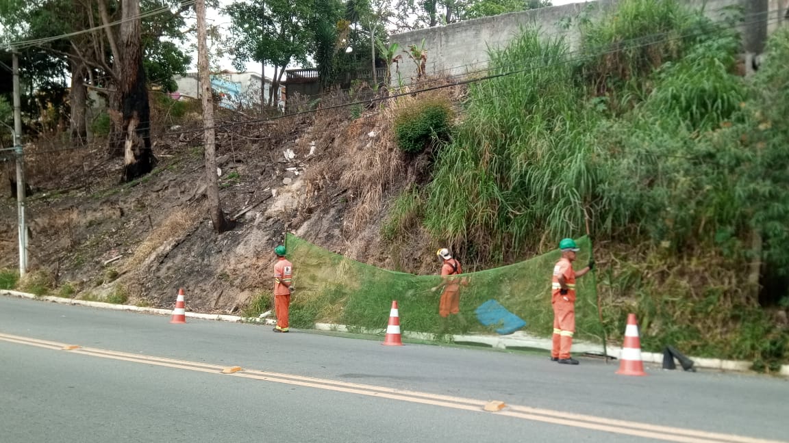 #PraCegoVer - Trabalhadores da Subprefeitura cortam grama na beira da via. Outros seguram rede de proteção. Atrás do local, há uma encosta.