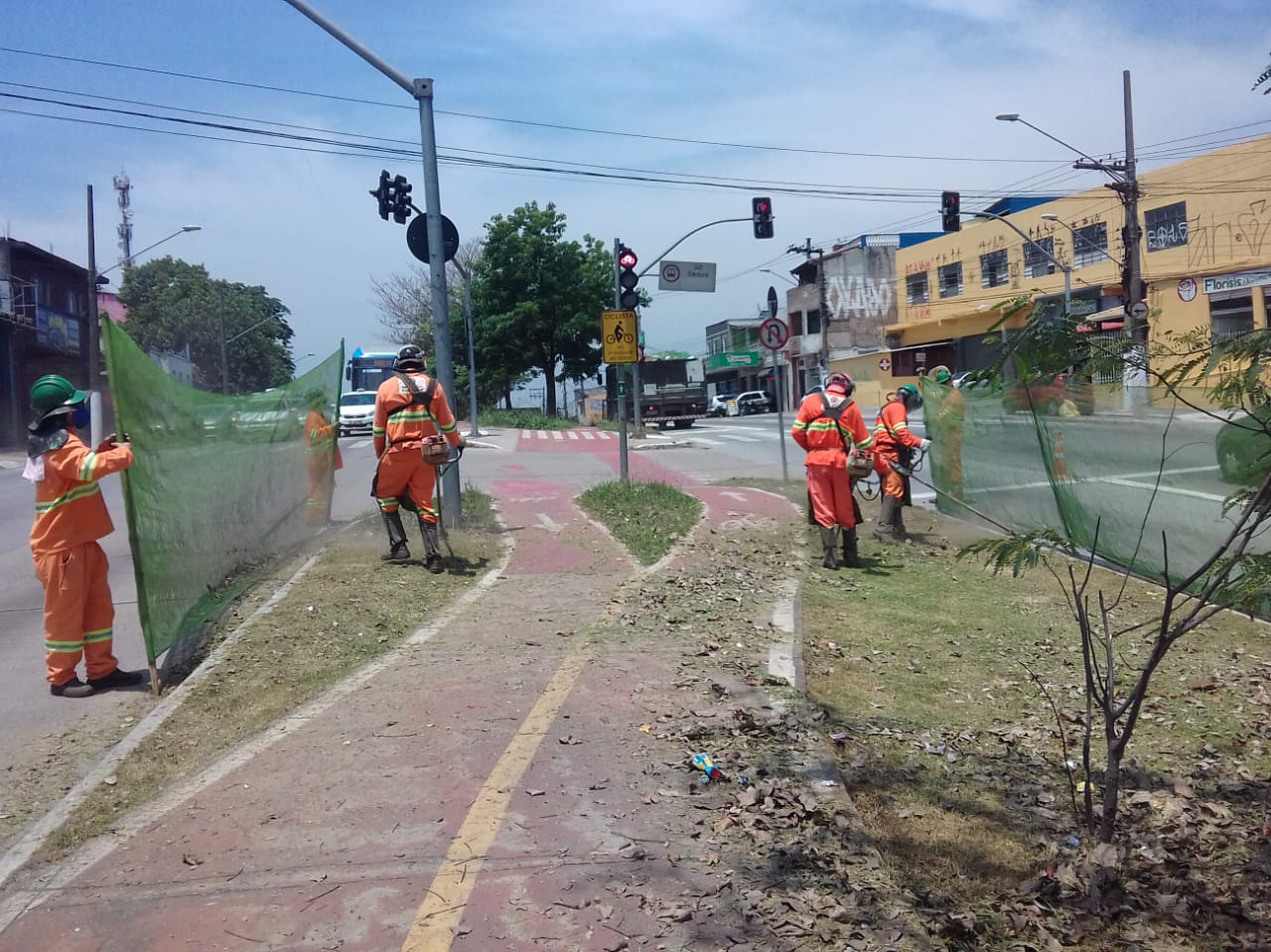 #PraCegoVer - Trabalhadores da Subprefeitura cortam grama no canteiro central. Outros seguram proteção de tela. No canteiro central, há uma ciclovia. Do lado direito da via, há um prédio cor de creme forte, quase amarelo.