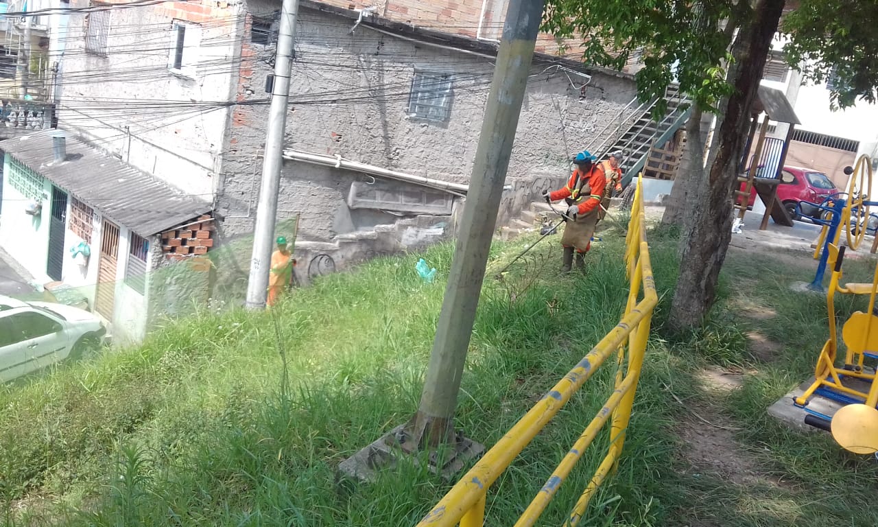 #PraCegoVer - Trabalhadores da Subprefeitura cortam grama ao lado de um guarda-corpo pintado de amarelo, ao lado de uma encosta. Outros trabalhadores seguram a proteção de tela. À direita, há aparelhos de ginástica de terceira idade. No fundo, à esquerda, há uma casa com a parede rebocada, mas não pintada.