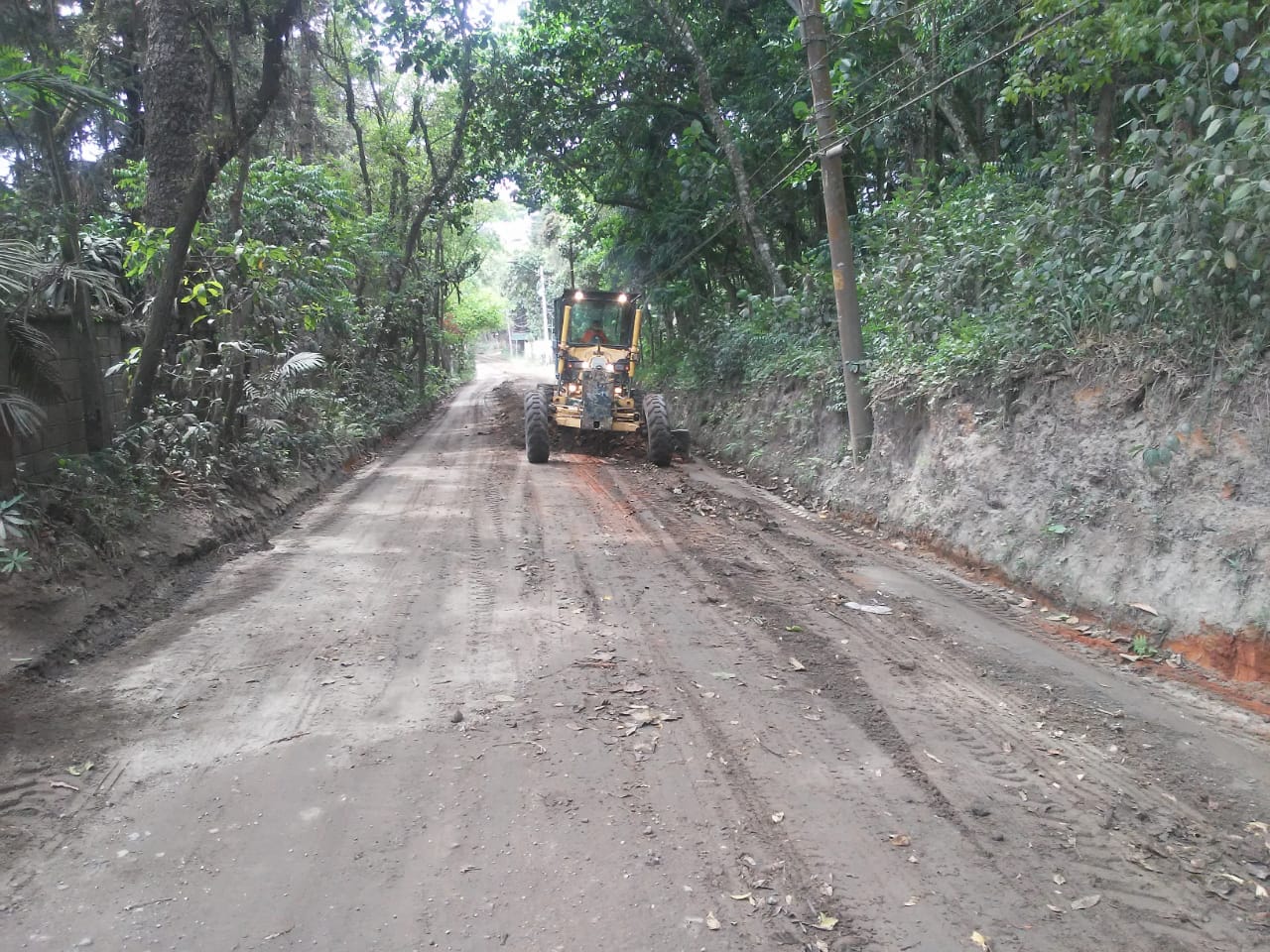 #PraCegoVer - Motoniveladora nivela a via de terra. Dos dois lados da via, há vegetação. Do lado direito, também postes.