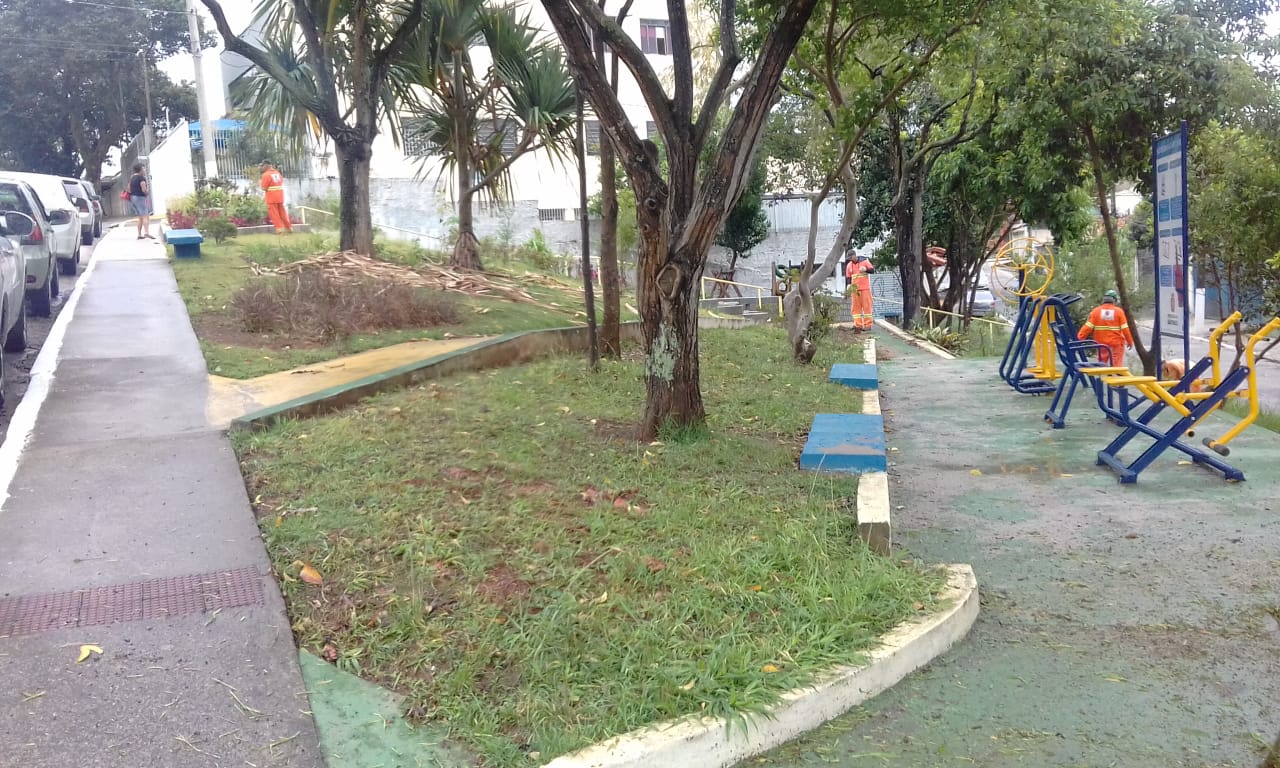 #PraCegoVer - Trabalhadores da Subprefeitura cortam grama na praça. Do lado direito, há aparelhos de ginástica.