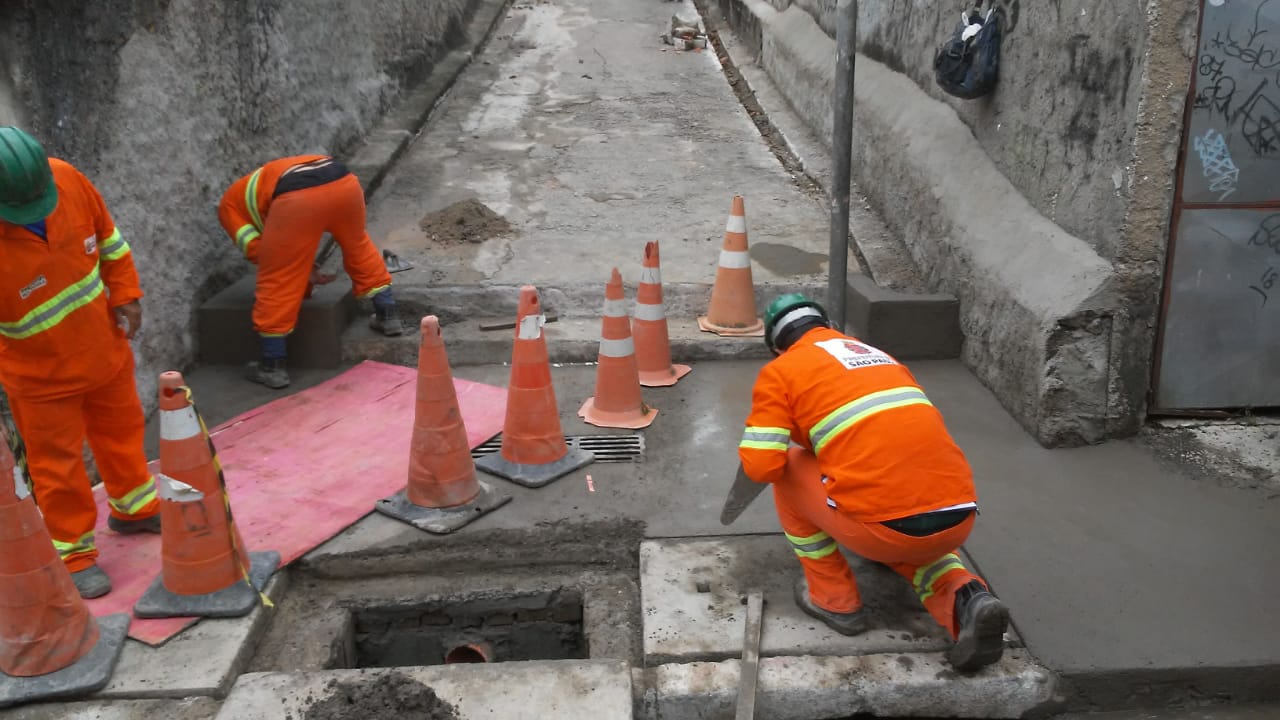 #PraCegoVer - Trabalhadores da Subprefeitura fazem arremate na parte baixa da viela. A galeria de águas pluviais (GAP) está com uma das tampas aberta. O local está sinalizado.