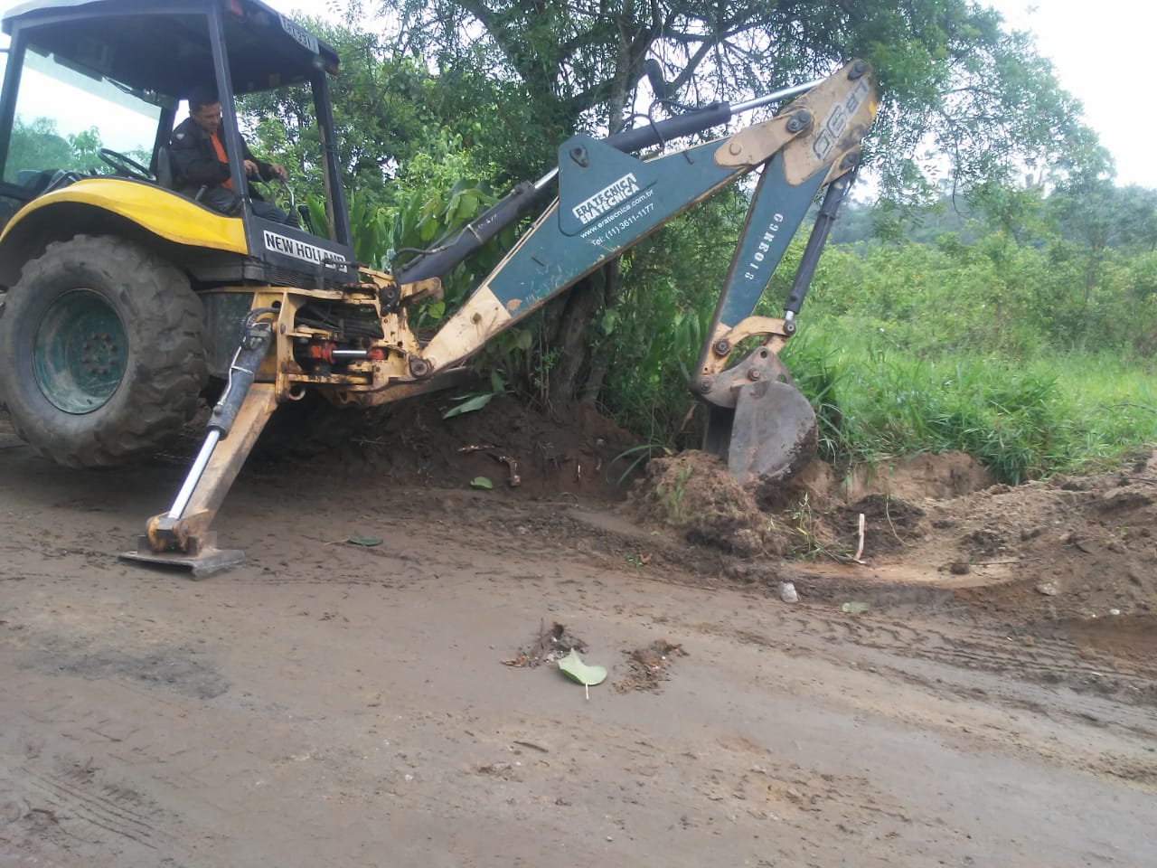 #PraCegoVer - Escavadeira remove terra da margem da via, para abrir canal de escoamento de água. As margens são cobertas por vegetação.