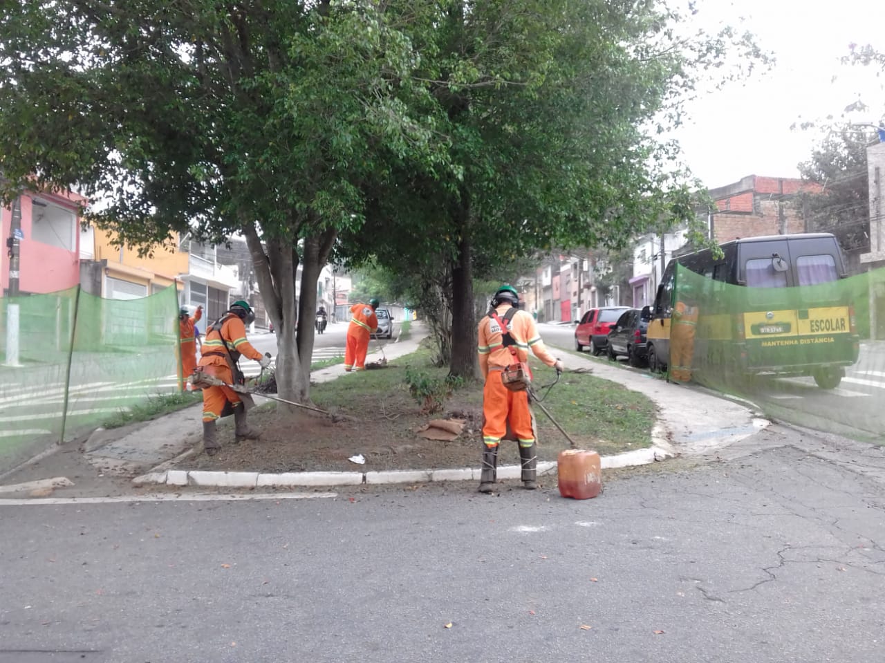#PraCegoVer - Trabalhadores da Subprefeitura cortam grama no canteiro central da avenida, coberto por árvores. Outros trabalhadores seguram proteções de tela.