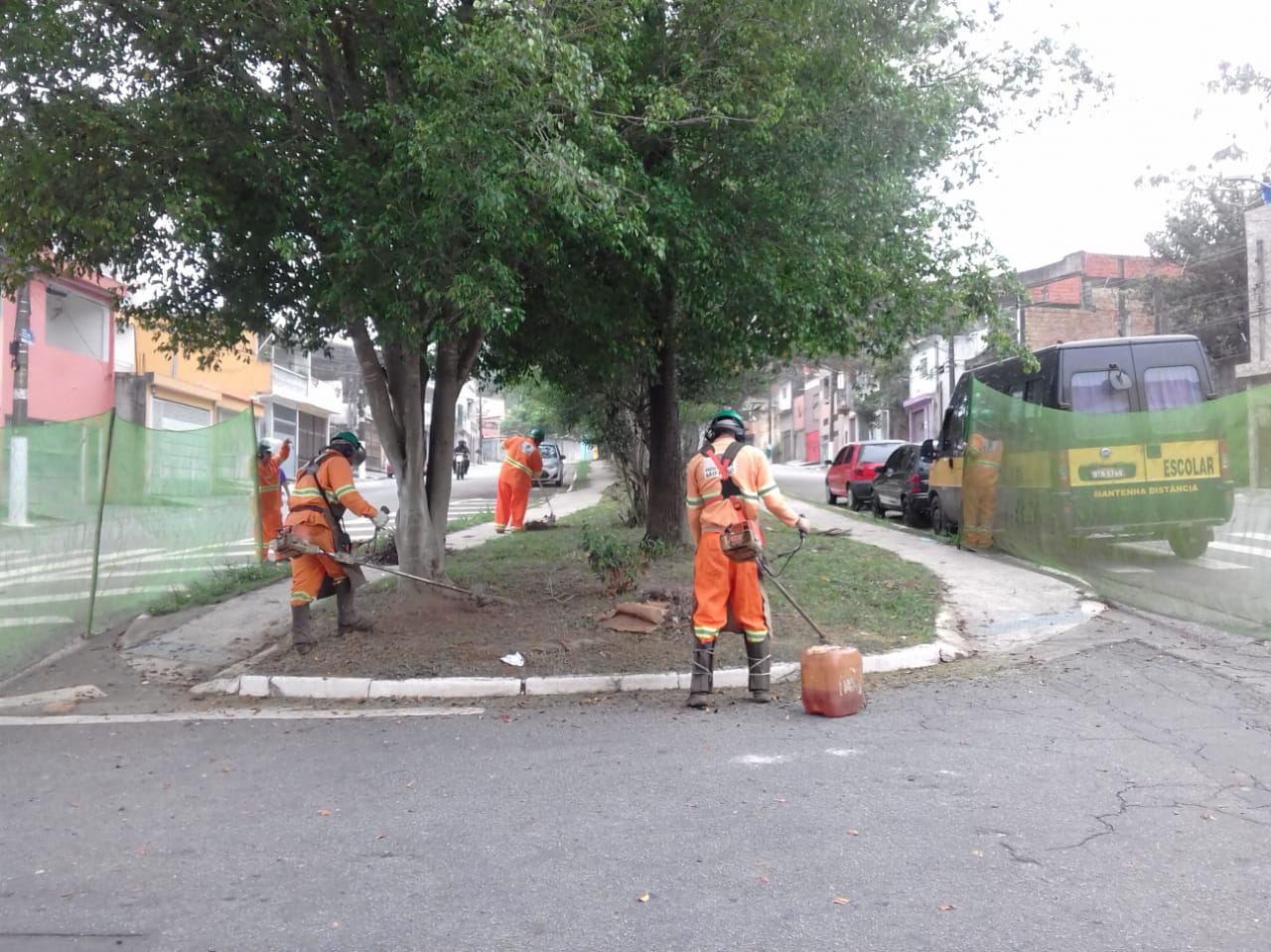 #PraCegoVer - Trabalhadores da Subprefeitura cortam grama no canteiro central da avenida, coberto por árvores. Outros trabalhadores seguram proteções de tela.