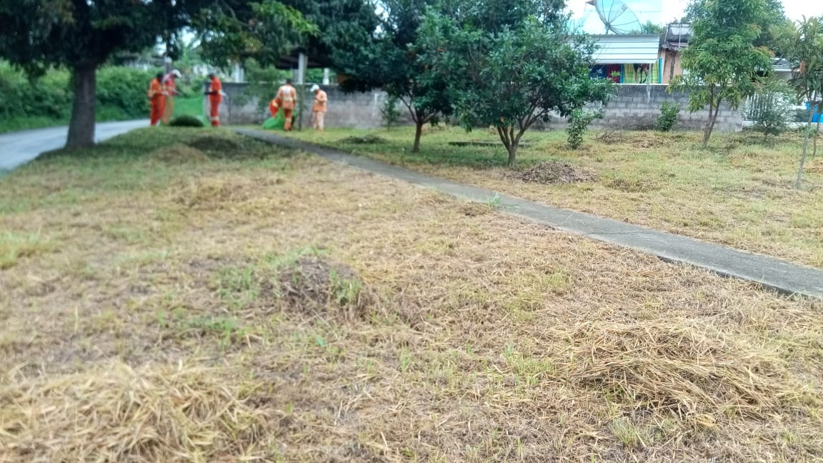 #PraCegoVer - Ao fundo, trabalhadores da Subprefeitura fazem corte de grama na praça. Em primeiro plano, a grama anteriormente cortada. O local tem árvores e um passeio transversal.