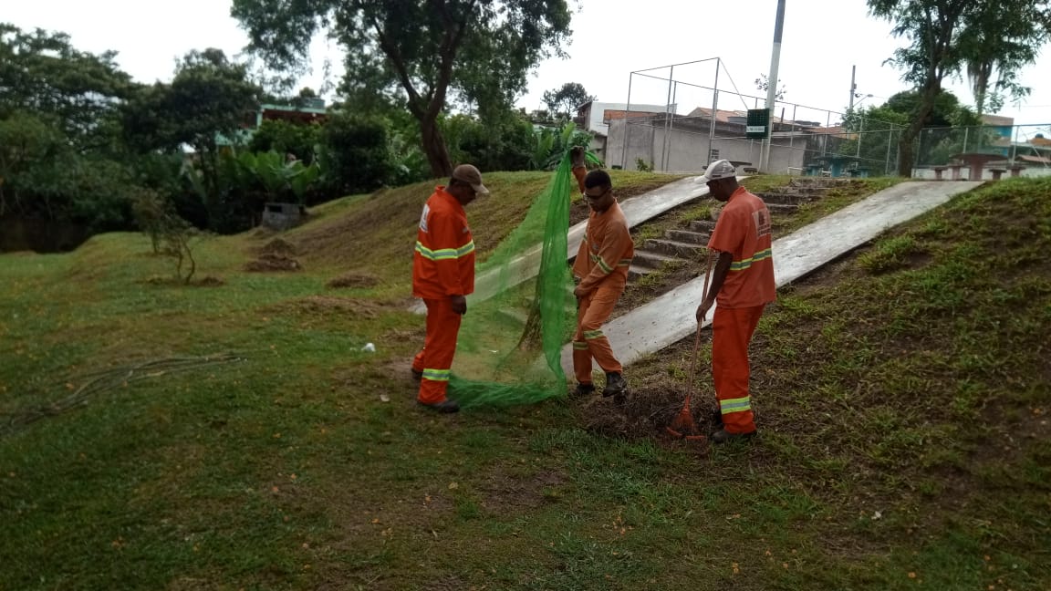 #PraCegoVer - Trabalhadores da Subprefeitura recolhem grama e galhos cortados na praça. Ao fundo, há uma escadaria e dois escorregadores de cimento. Mais ao fundo, uma quadra de esportes. Tudo em meio a árvores.