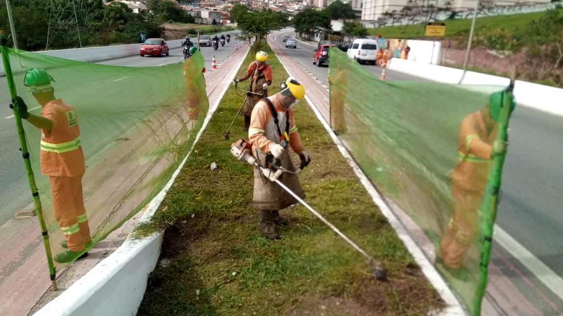 #PraCegoVer - Dois trabalhadores da Subprefeitura cortam grama no canteiro central. Outros quadro seguram proteções de tela dos dois lados do canteiro.