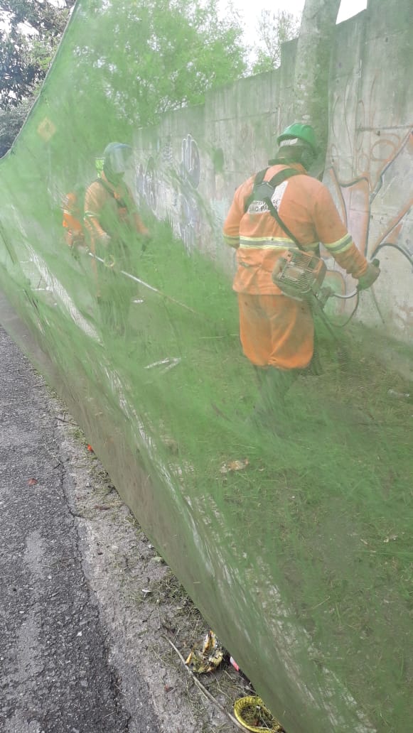 #PraCegoVer - Dois trabalhadores da Subprefeitura cortam grama por trás de uma proteção de tela. Eles estão diante de um muro de placas de cimento.