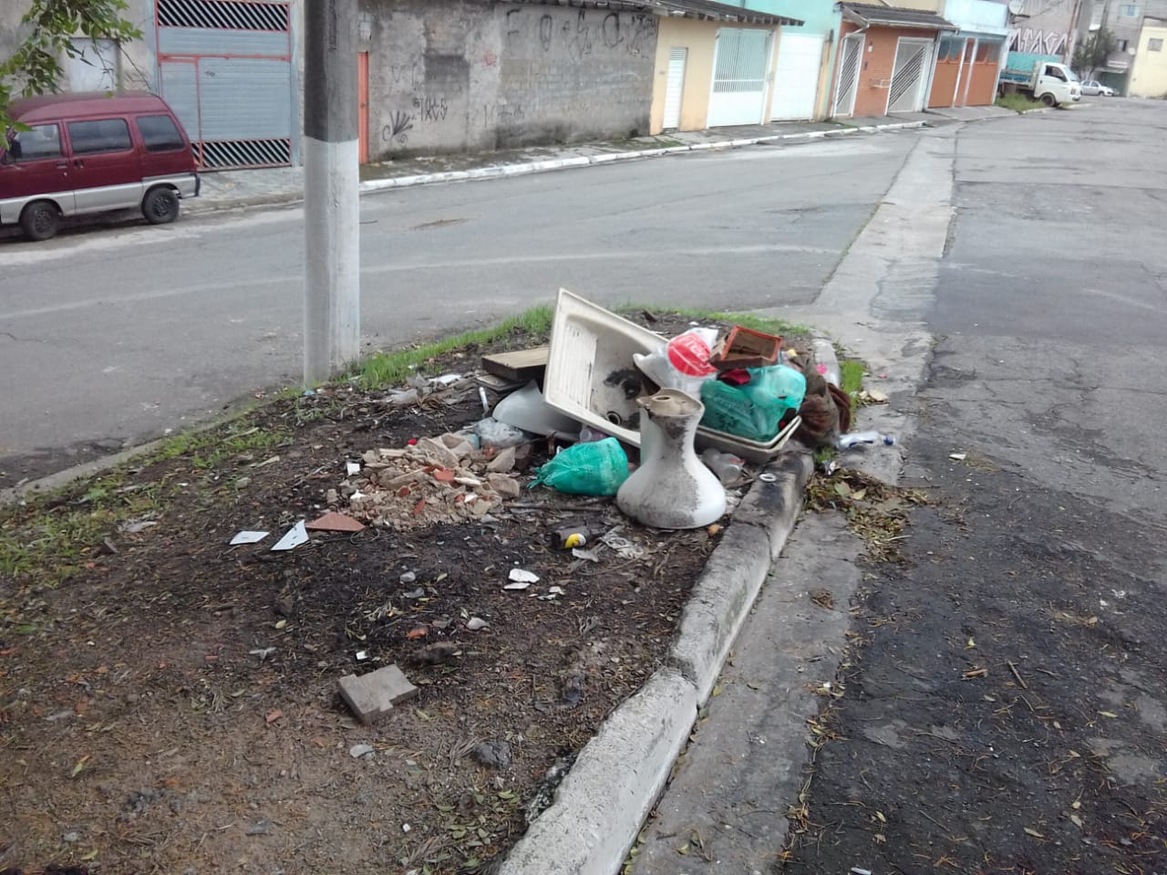 #PraCegoVer - Lixo e entulho no bico de um canteiro no encontro de duas ruas. Entre os entulhos há uma bacia sanitária e um tanque de lavar roupas.