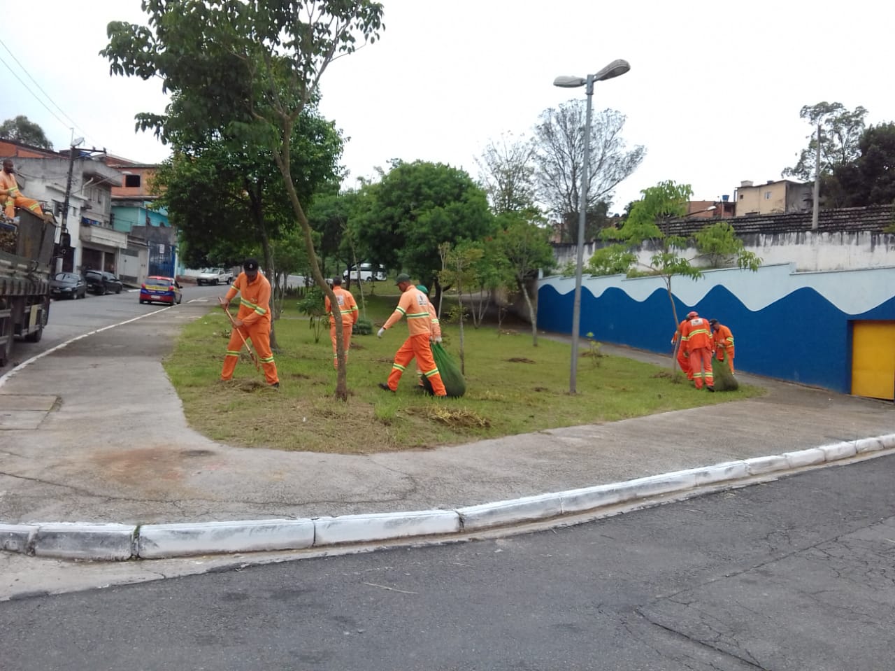 #PraCegoVer - Trabalhadores da Subprefeitura recolhem grama e galhos do corte em espaço gramado em esquina. Há árvores no local. E, do lado direito, um muro pintado de azul e branco em ondas.