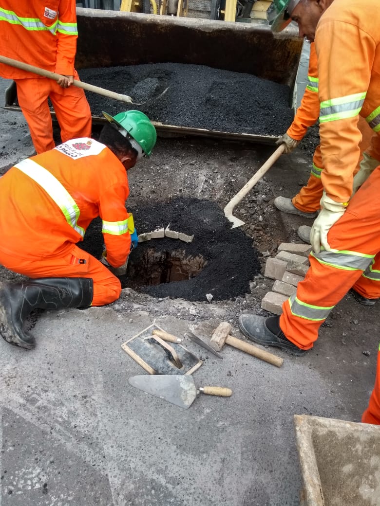 #PraCegoVer - Trabalhadores da Subprefeitura expuseram o poço-de-visita (PV) para levantá-lo ao nível do leito. Alguns homens colocam asfalto nas beiradas. Pelo chão, há ferramentas, como desempenadeira, colher de pedreiro e marreta.