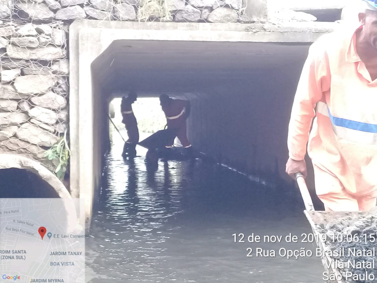 #PraCegoVer - Dois trabalhadores da Subprefeitura estão dentro de uma aduela (peça de concreto no formato de um túnel quadrado), recolhendo sujeira do córrego. Em primeiro plano, um trabalhador leva, em carrinho-de-mão, detritos recolhidos.