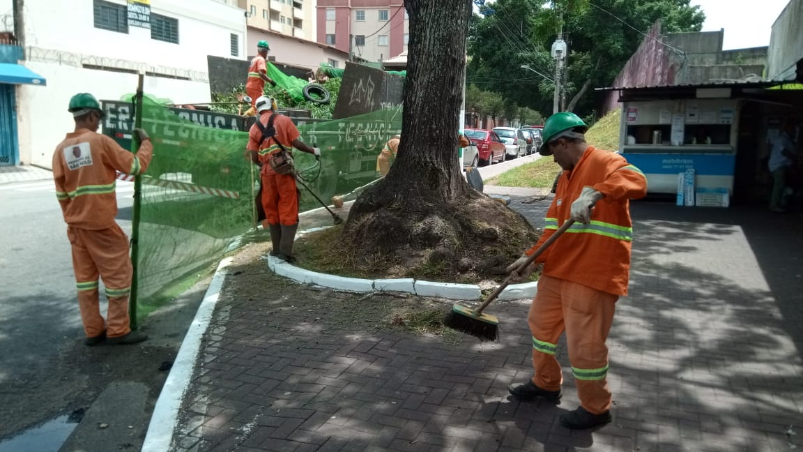 #PraCegoVer - Trabalhadores da Subprefeitura cortam grama em volta de uma árvore. Outros recolhem a grama cortada em um caminhão da empreiteira. E um outro, ainda, varre o local.