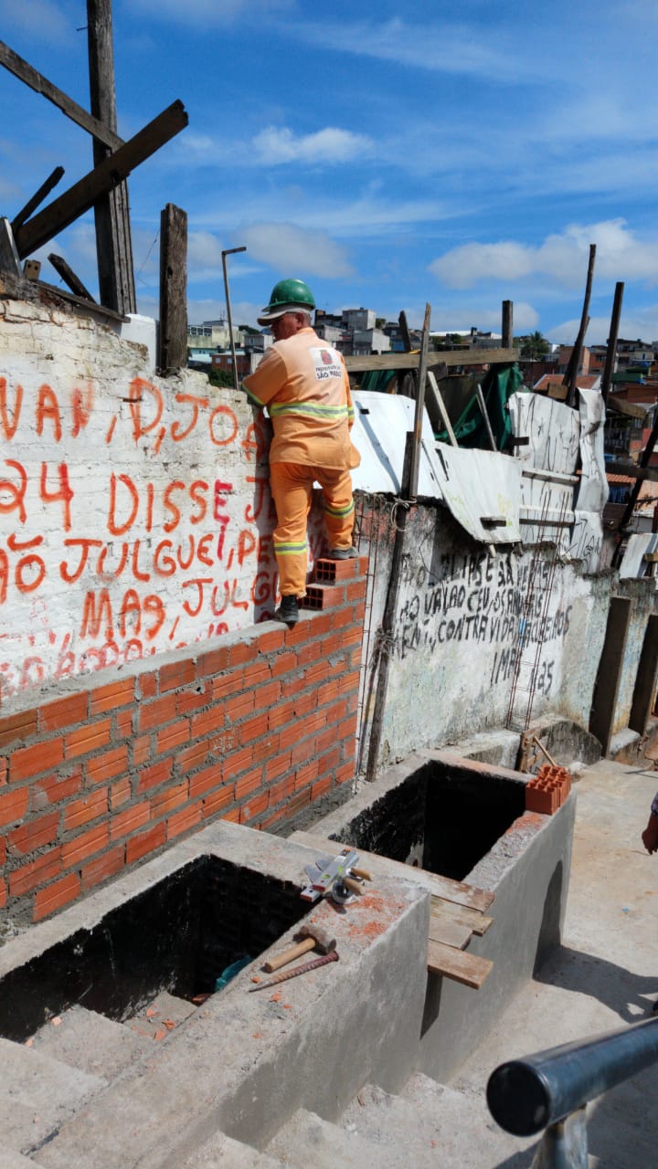 #PraCegoVer - Trabalhador da Subprefeitura está sobre um muro que está sendo construído. Na parte inferior, há degraus e duas caixas de concreto.