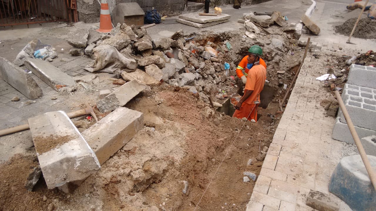 #PraCegoVer - Trabalhadores da Subprefeitura escavam o local da boca de lobo. Paralelepípedos estão amontoados do lado esquerdo, assim como entulho resultante da escavação. Do lado direito, blocos de concreto.
