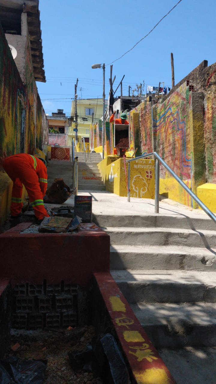 #PraCegoVer - Trabalhadores da Subprefeitura arrematam com cimento as construções de caixas na viela. As caixas estão pintadas de vermelho ou amarelo.