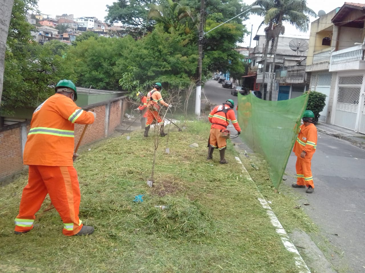 #PraCegoVer - Trabalhadores da Subprefeitura cortam grama no passeio da via. Outros seguram a proteção de tela.