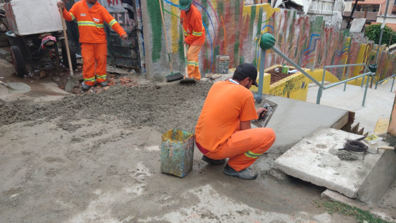 #PraCegoVer - Trabalhador da Subprefeitura pinta o corrimão da viela. Outros trabalhadores espalham cimento na plataforma inicial. Os muros estão pintados de cores vivas. A caixa de contenção da água de chuva está pintada de amarelo.