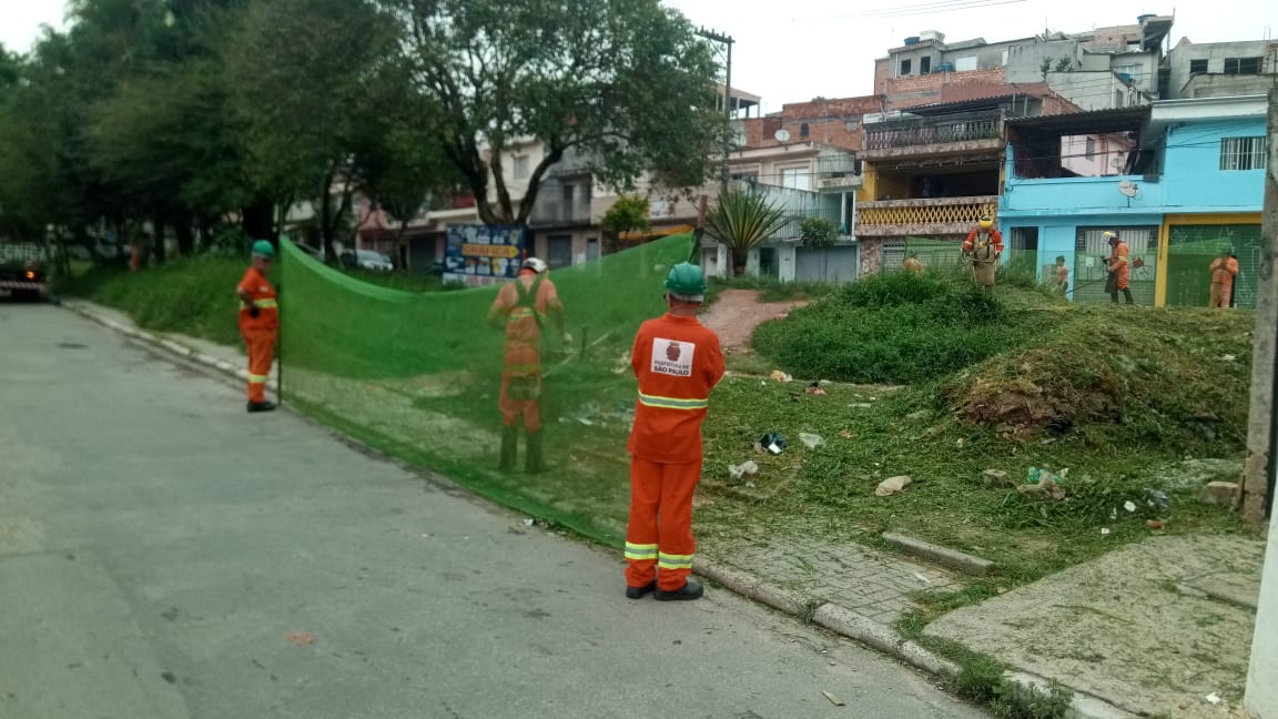 #PraCegoVer - Trabalhadores da Subprefeitura cortam grama em dois níveis do jardim entre duas vias. Outros seguram proteções de tela.