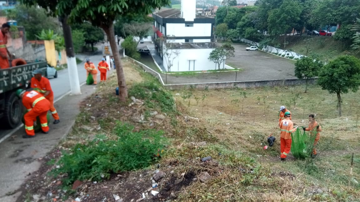 #PraCegoVer - Trabalhadores da Subprefeitura recolhem grama cortada. A rua tem um jardim em desnível para o lado direito. Há árvores em quase todo o espaço.