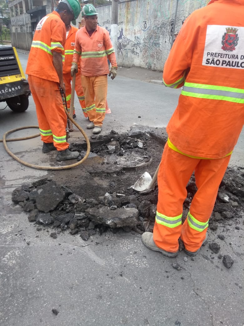 #PraCegoVer - Trabalhadores da Subprefeitura retiram o asfalto em volta do poço-de-visita (bueiro com tampa) que será consertado.