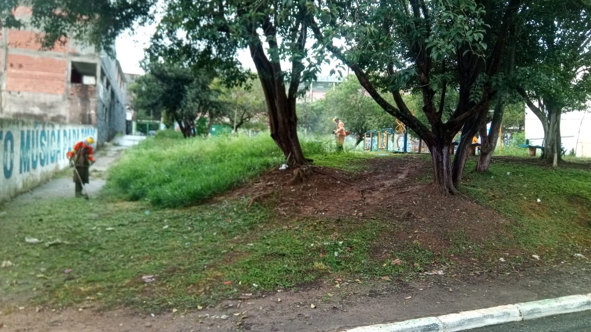 #PraCegoVer - Trabalhaodres da Subprefeitura cortam grama em dois pontos de espaço aberto na via. Há árvores no terreno.