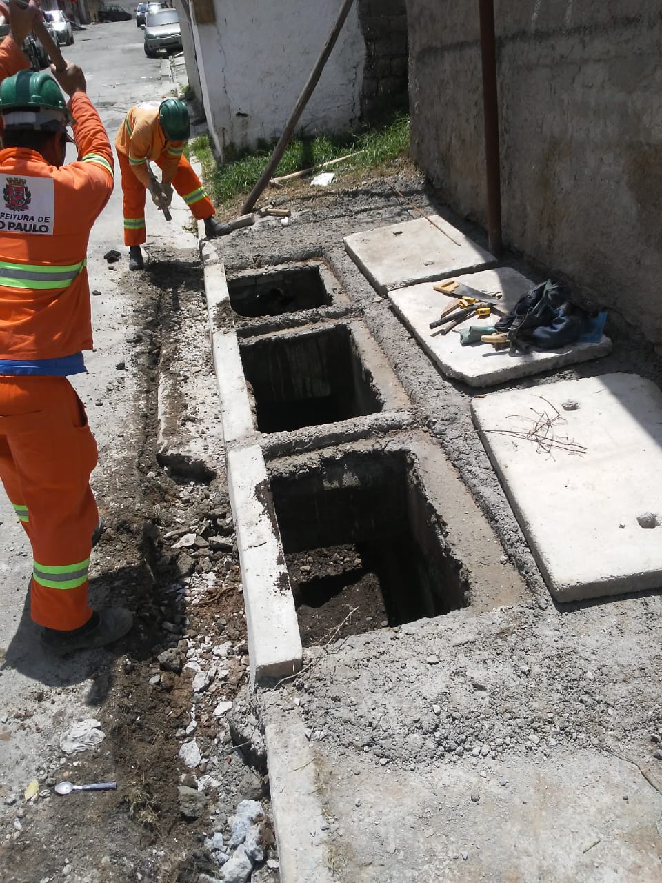 #PraCegoVer - Trabalhadores da Subprefeitura estão diante de três bocas-de-lobo (BLs), procedendo ao conserto. As três BLs estão abertas, com as tampas colocadas no passeio, contra um muro.