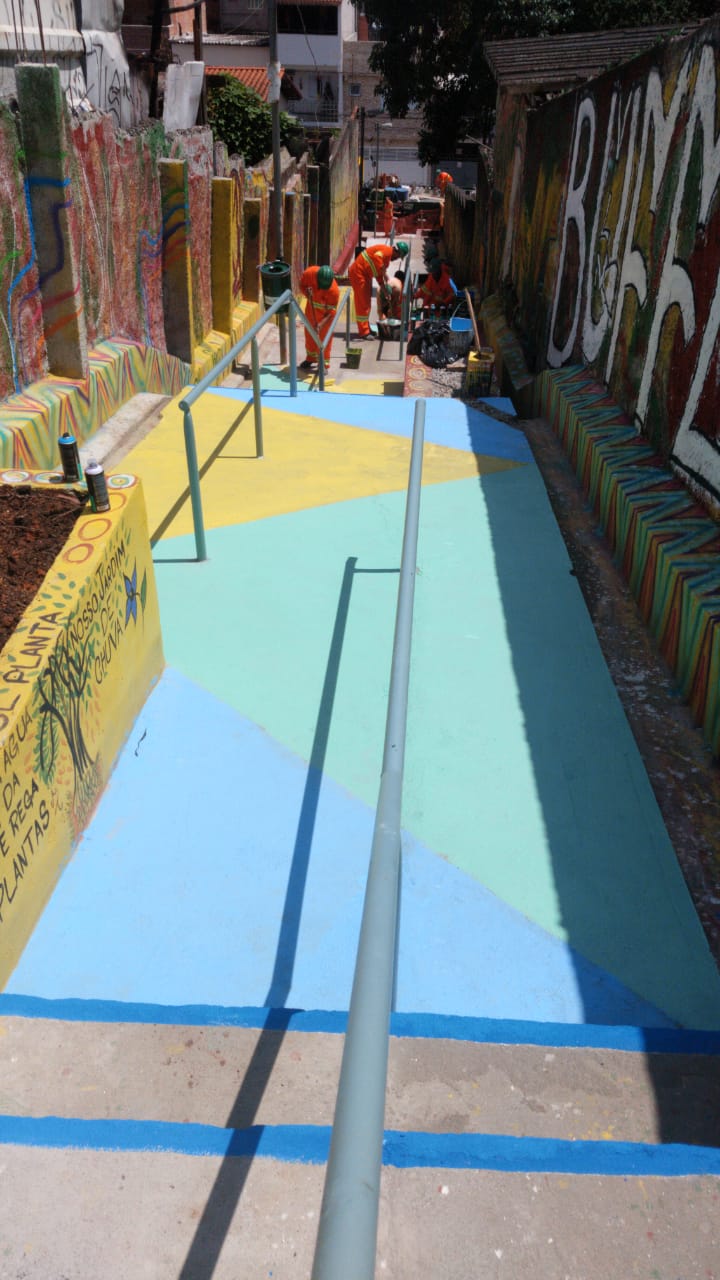 #PraCegoVer - Trabalhadores da Subprefeitura pintam o piso do escadão com desenhos geométricos e coloridos. Há caixas de retenção de água da chuva, pintadas de amarelo e vermelho. Um corrimão atravessa todo o escadão, de cima a baixo.
