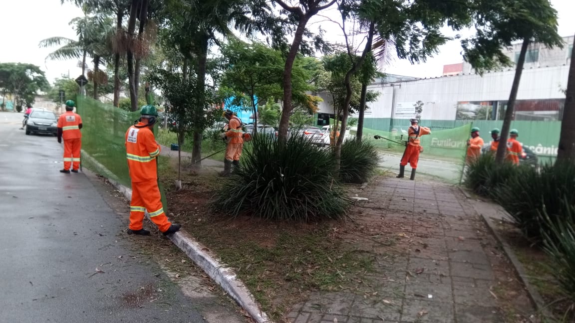 #PraCegoVer - Trabalhadores da Subprefeitura cortam grama e recolhem galhos jogados na calçada central. Outros trabalhadores seguram uma proteção de tela.