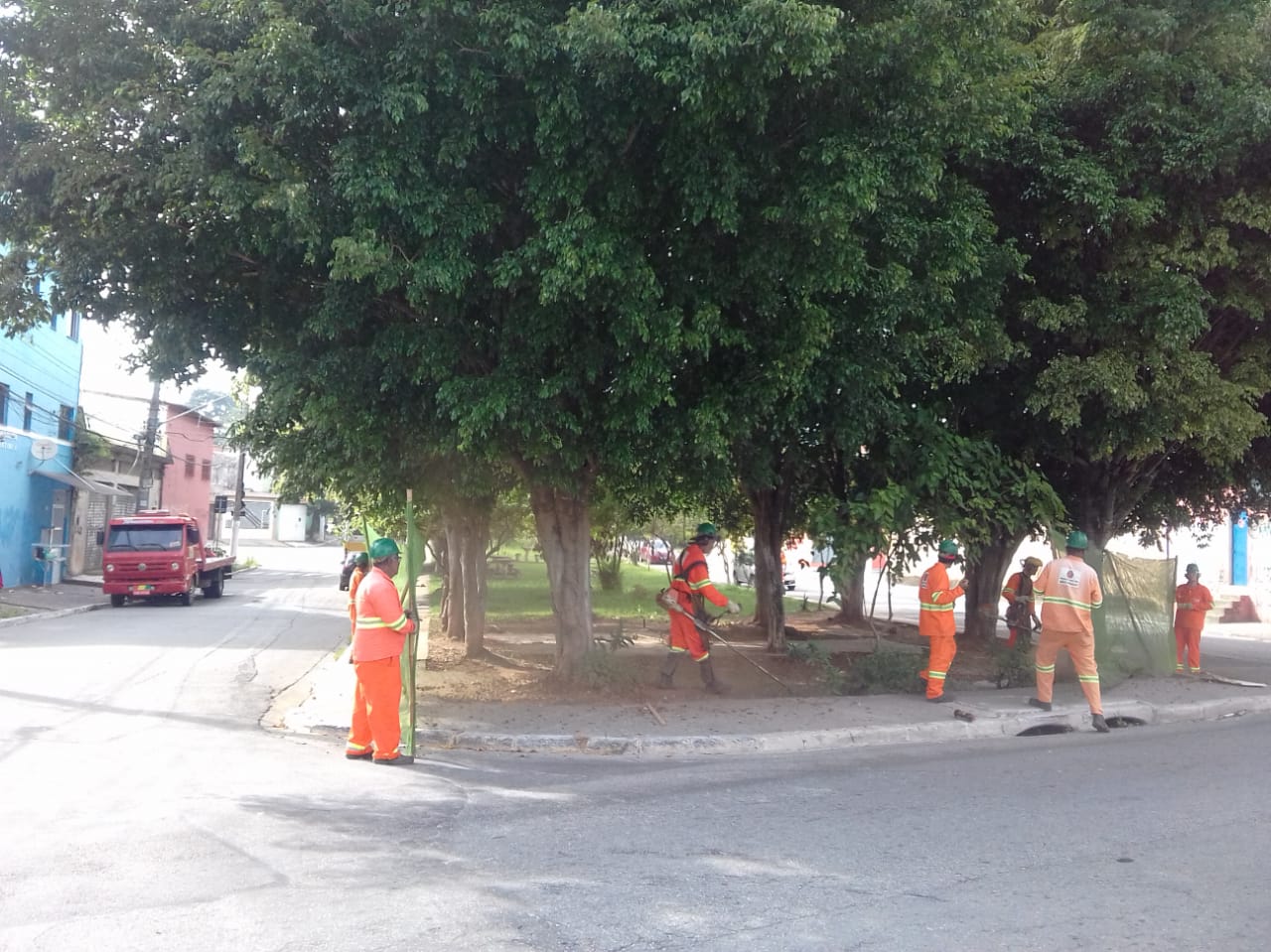#PraCegoVer - Trabalhadores da Subprefeitura cortam grama no jardim da via. Outros seguram proteção de tela. O local tem árvores bem frondosas.
