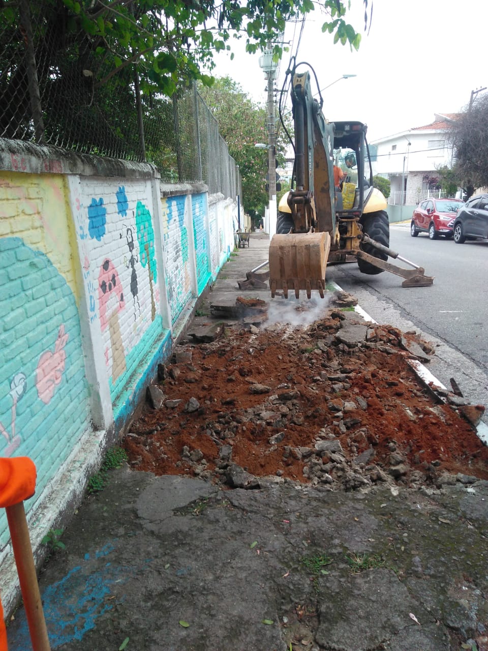 #PraCegoVer - Uma escavadeira quebra o passeio que será reformado. Ao lado, há um muro. E carros ao fundo.
