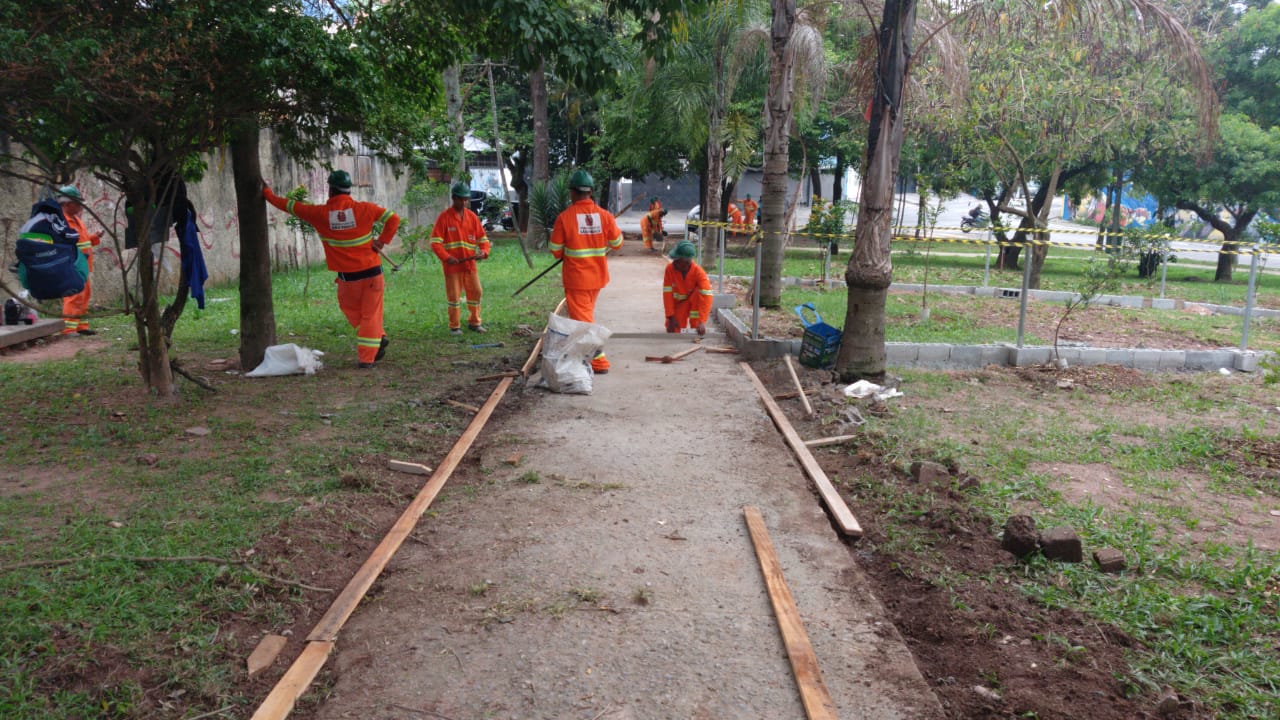 #PraCegoVer - Trabalhadores da Subprefeitura fazem obras na calçada da praça. O caminho está delimitado por ripas de madeira. Há árvores e gramado. 