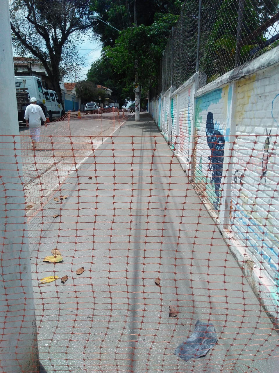 #PraCegoVer - Passeio já cimentado em frente a muro pintado com desenhos. O local está protegido com uma tela de plástico, que impede a passagem de pedestres, até o cimento secar.