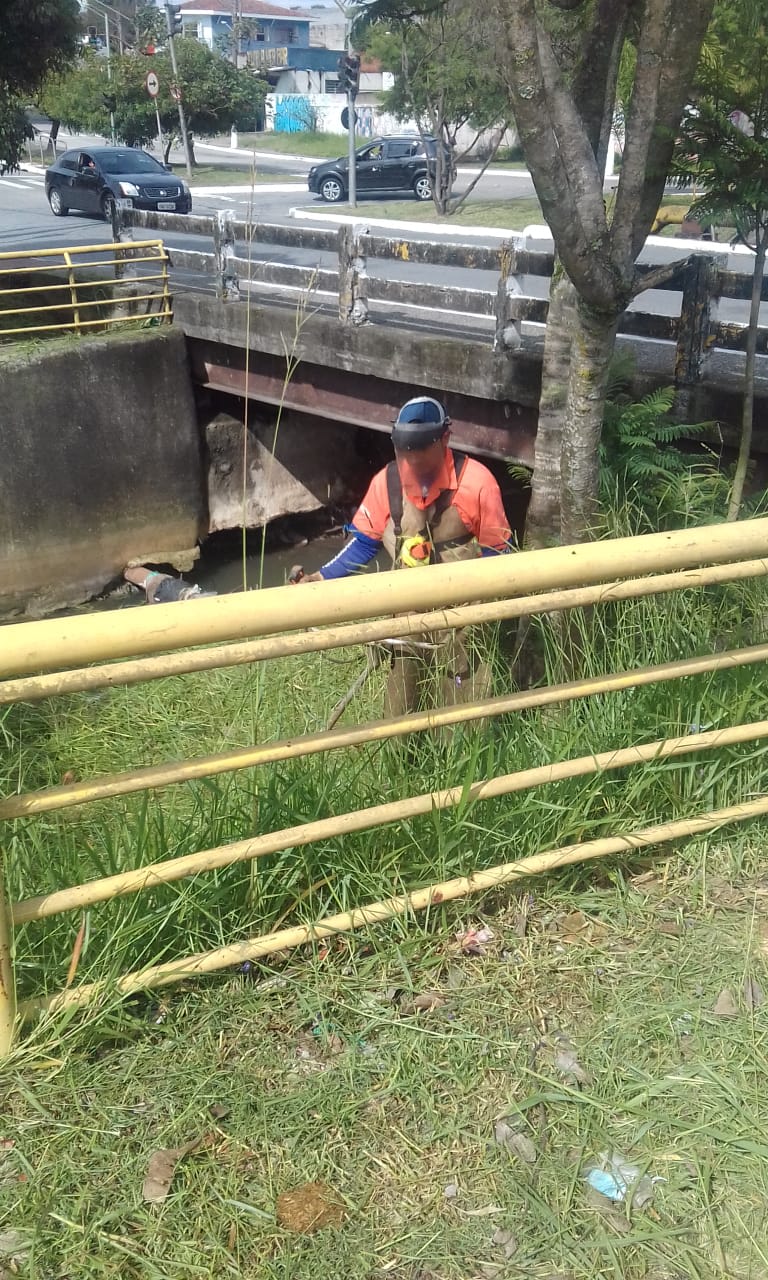#PraCegoVer - Trabalhador da Subprefeitura corta mato e grama por trás de um guarda-corpo de ferro pintado de amarelo.