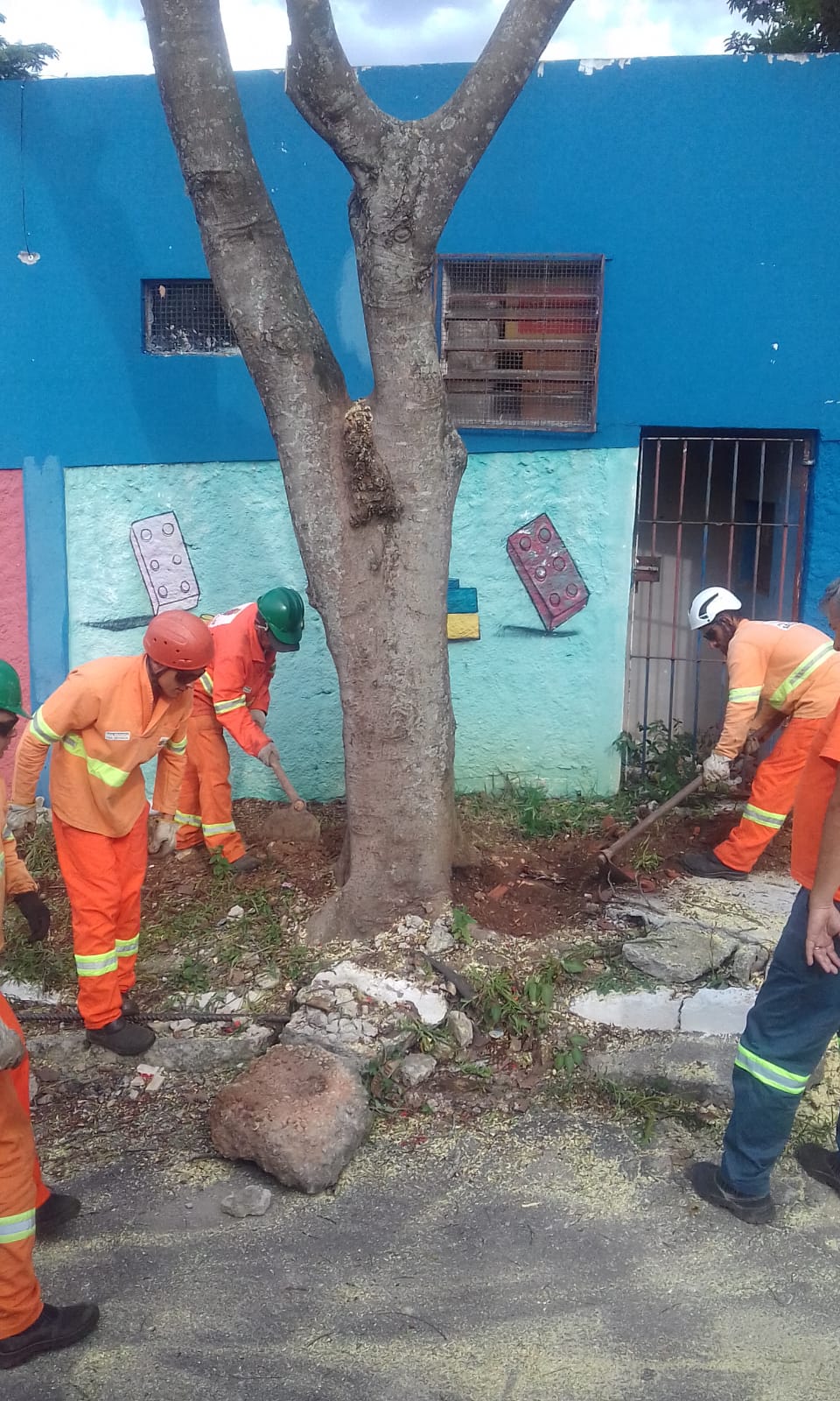#PraCegoVer - Trabalhadores da Subprefeitura escavam em volta de árvore que será removida. Ao fundo há um prédio azul escuro com desenho pintado em azul claro.