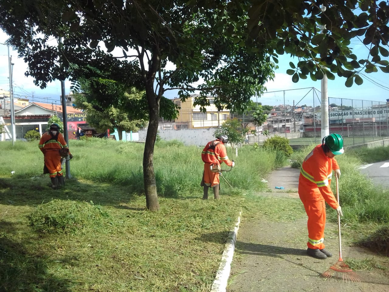 #PraCegoVer - Trabalhadores da Subprefeitura cortam grama no passeio, ao lado de uma quadra de esportes.