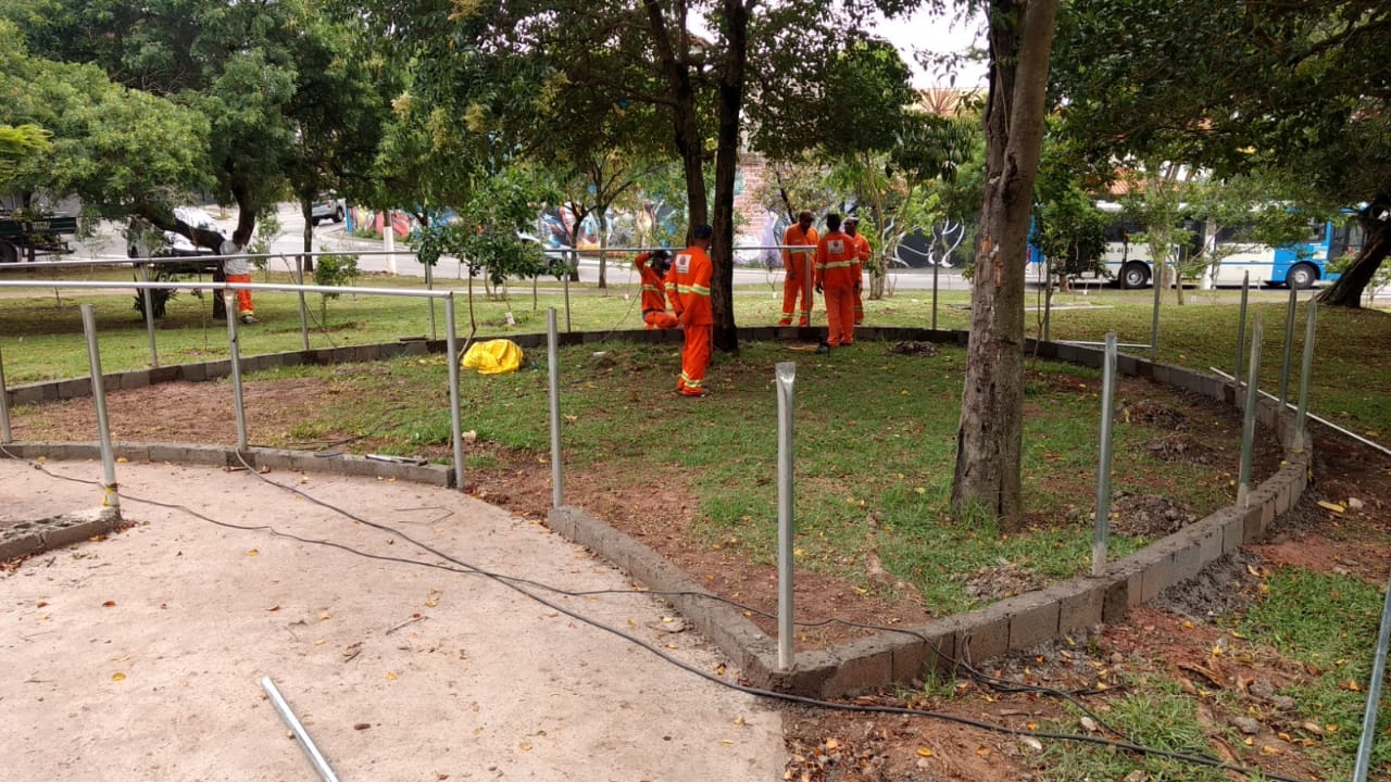 #PraCegoVer - Trabalhadores da Subprefeitura montam uma cerca de metal na praça. Há árvores e a grama está aparada.