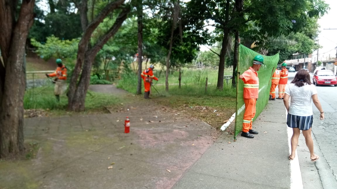 #PraCegoVer - Trabalhadores da Subprefeitura cortam a grama de um pequeno bosque ao lado da calçada. Outros trabalhadores seguram proteção de tela.