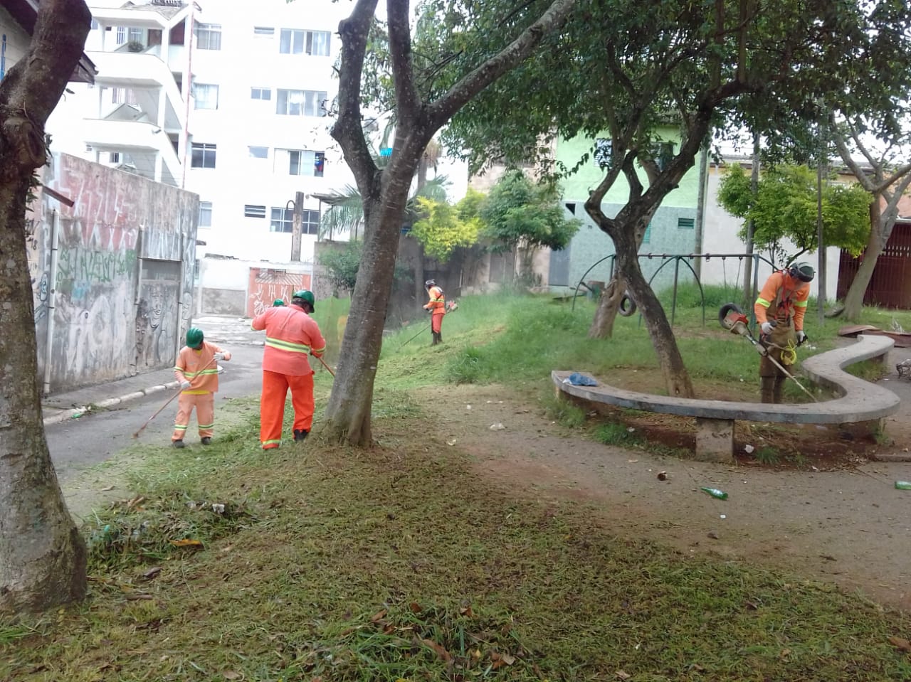 #PraCegoVer - Trabalhadores da Subprefeitura cortam grama em praceta do local. Há um banco de concreto de formato curvo. Há árvores no local.