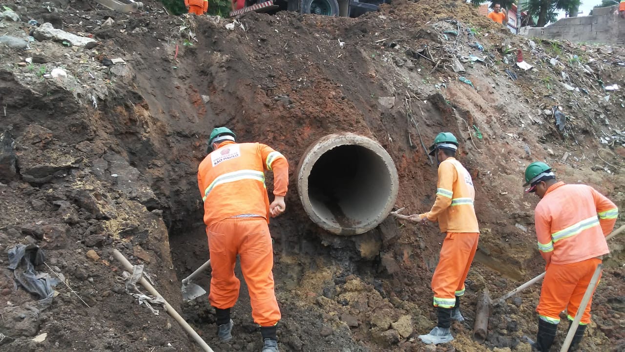 #PraCegoVer - Trabalhadores da Subprefeitura cavam terra em volta de tubulação em uma encosta.